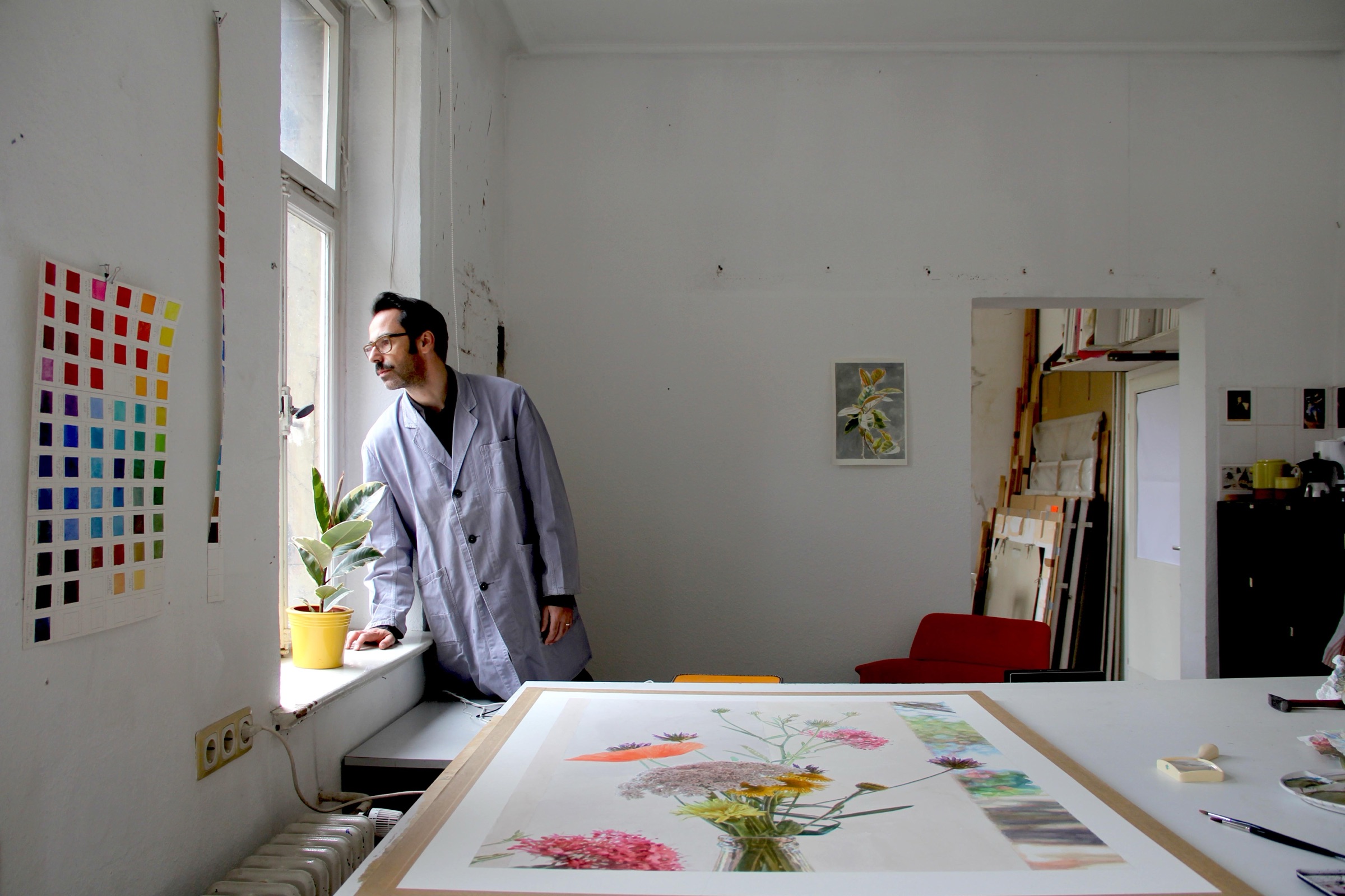 Peter Holl im hellen Atelier, er lehnt am Fenster und blickt hinaus. Im Vordergrund liegt ein großes Aquarell mit Blumen auf dem Tisch.