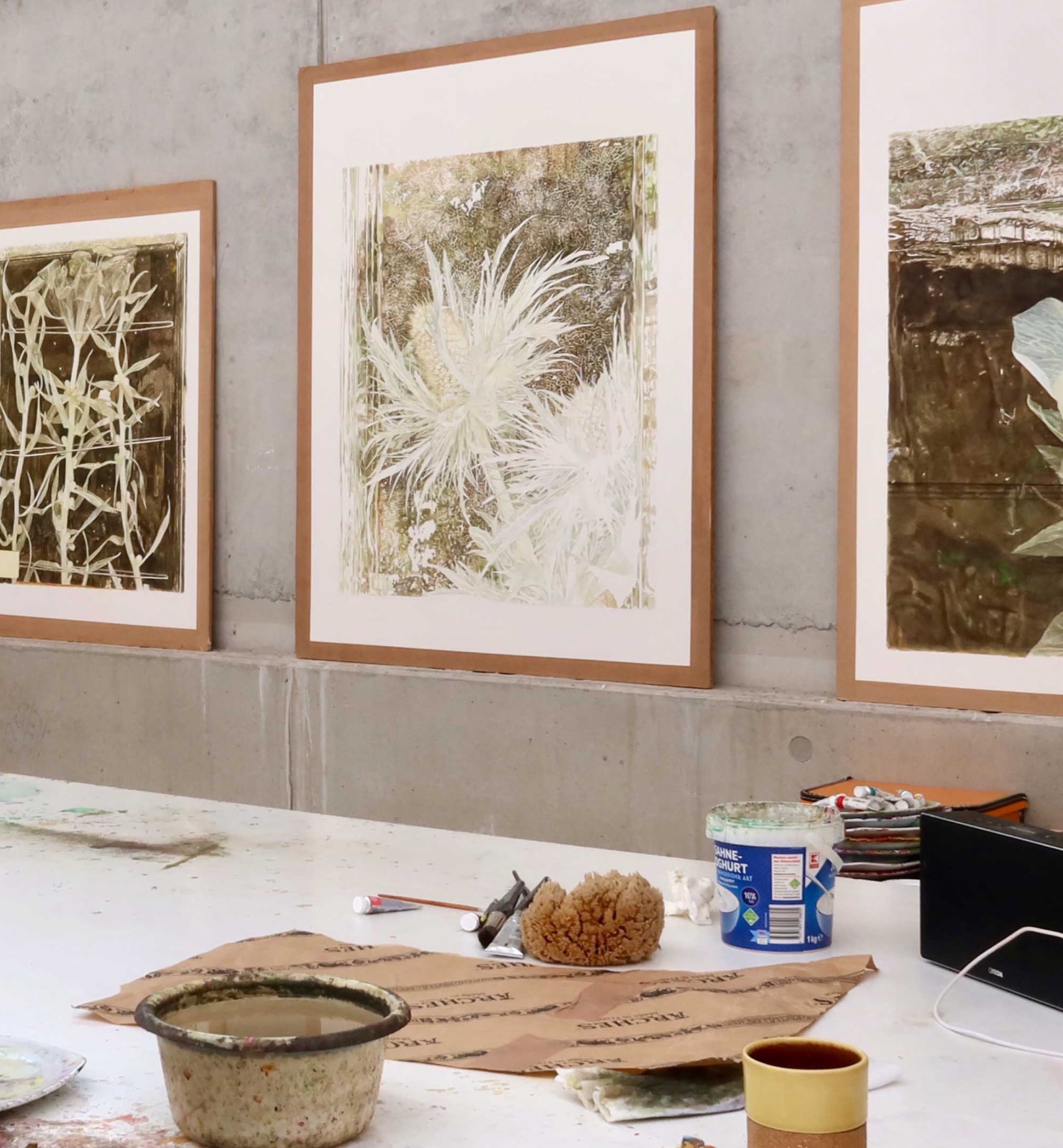 Blick in das Atelier: Drei große Aquarelle mit weißen Pflanzenmotiven auf dunklem Grund lehnen an einer Betonwand, davor ein Arbeitstisch mit Farbschale, Schwamm, Joghurtbecher und Tasse.