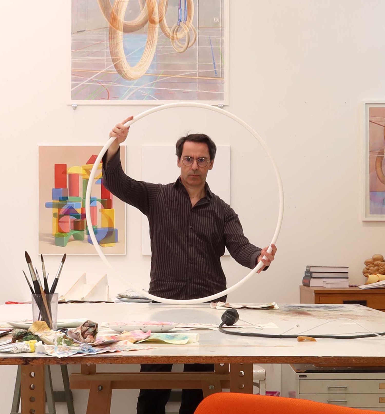 Peter Holl steht in seinem hellen Atelier hinter einem großen Arbeitstisch und hält einen weißen Reifen vor sich. An den Wänden hängen Aquarelle mit Holzbausteinen und Turnringen.