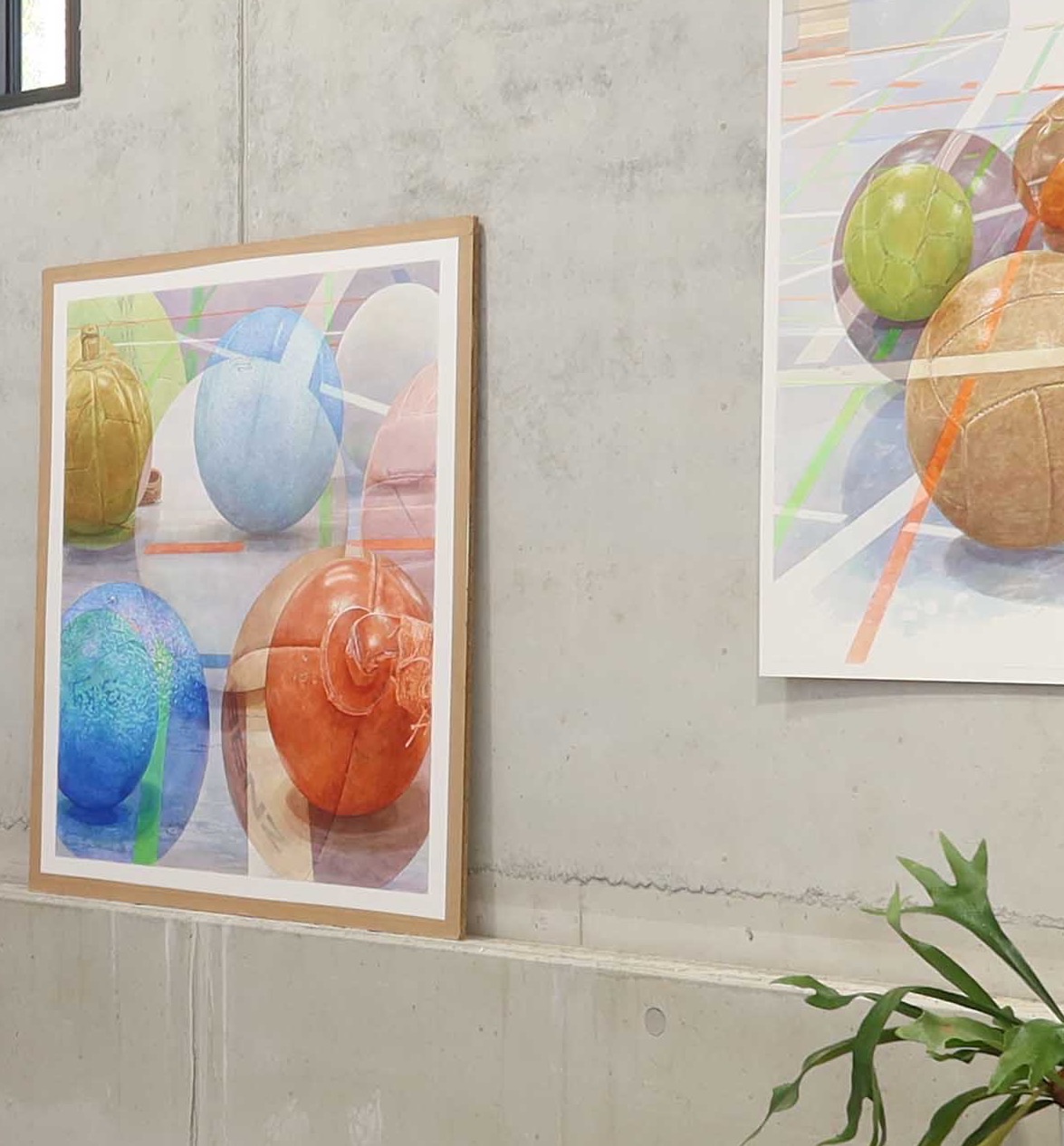 Blick in das Atelier: Ein gerahmtes Aquarell mit bunten Bällen lehnt an einer Betonwand, rechts hängt ein großes Aquarell mit Bällen auf Hallenboden, unten ragt die Pflanze eines Geweihfarns ins Bild.